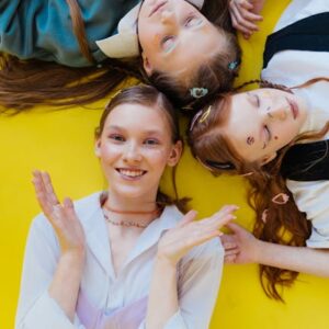 Tre smilende tenåringsjenter ligger med lukkede øyne på en livlig gul bakgrunn.